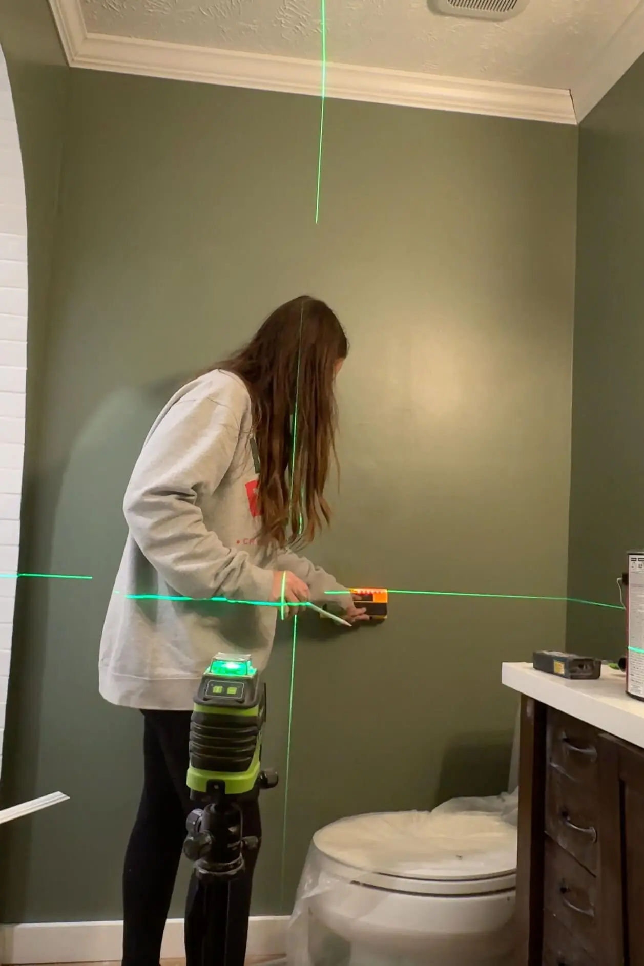 Person using a green laser level to mark precise measurements on a green bathroom wall during a DIY panel molding installation project.